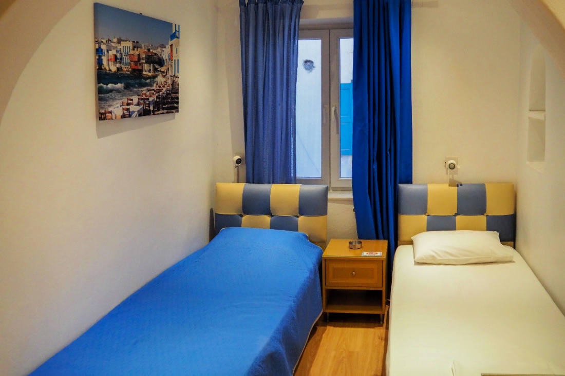 Zimmer in der günstigen Pension Ageliki Kalogera mit zwei Betten, blauen Vorhängen und Bild von Mykonos.