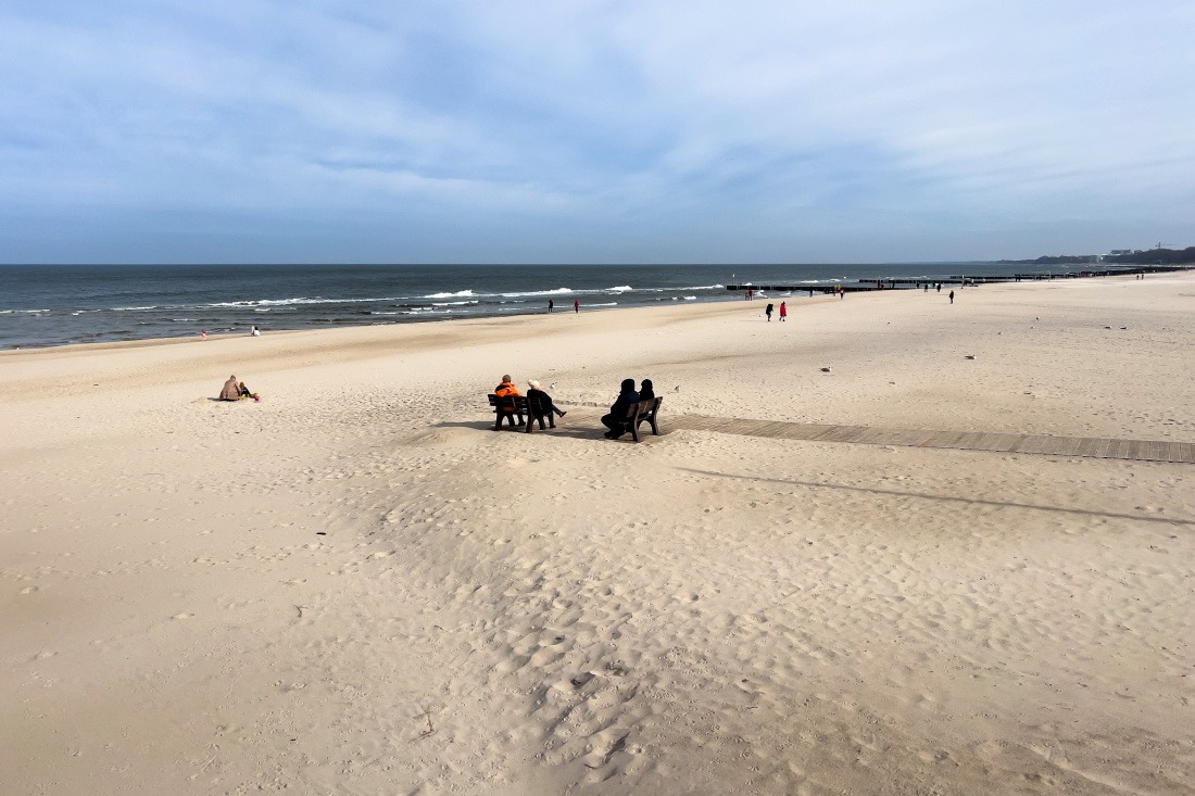 Der fast menschenleere Strand von Kolberg im März. Nur ein paar Menschen sitzen auf zwei Bänken oder laufen am Meer entlang.