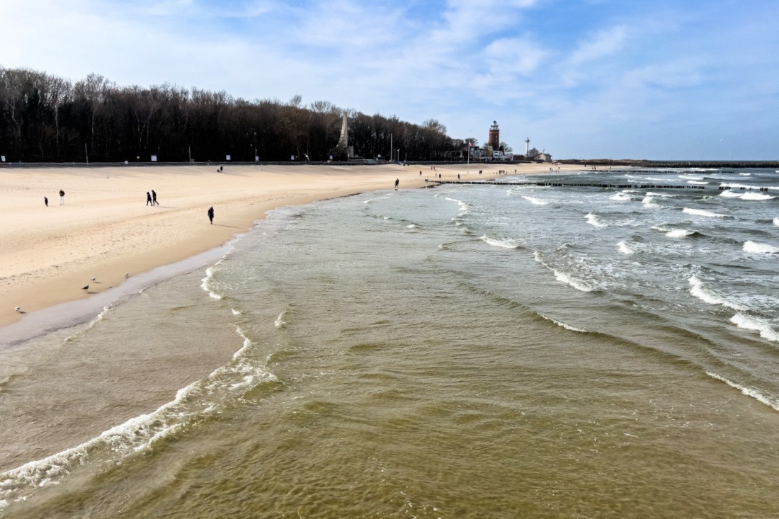 Blick von oben auf den Hauptstrand von Kolberg mit Leuchtturm in der Ferne. Auf der Ostsee kräuseln sich Wellen und am langen, breiten und goldfarbenen Strand laufen einzelne Menschen.