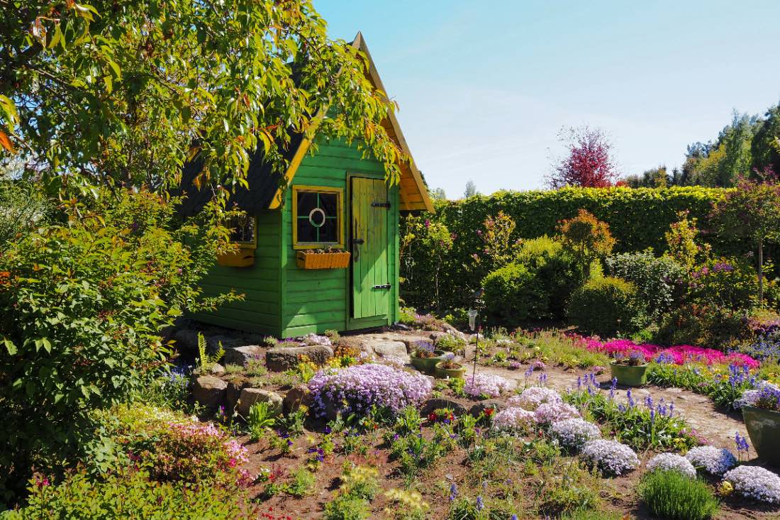 Die Hortulus Gärten liegen in der Umgebung von Kolberg. Das Bild zeigt eine grüne Hütte neben einem Baum, davor viele bunte Blumen. 