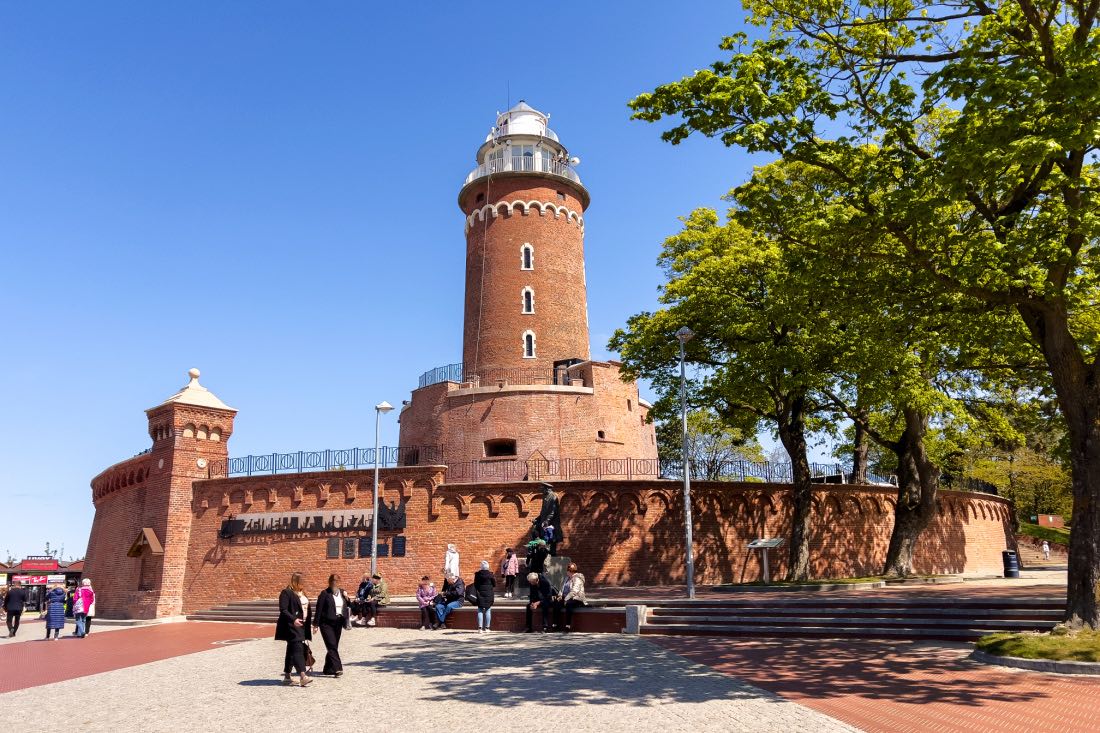 Zu den Kolberg Sehenswürdigkeiten zählt der Leuchtturm aus rotem Backstein. Er ist abgebildet vor blauem Himmel und mit einem großen Baum rechts daneben, davor laufen und sitzen Menschen. 