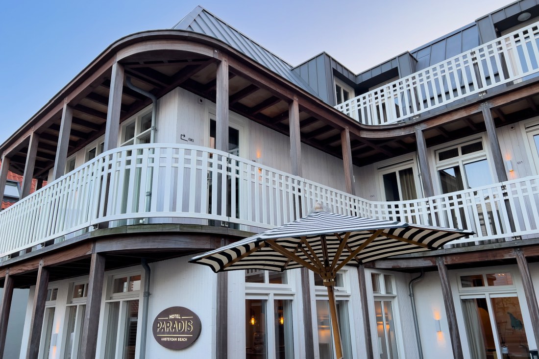 Das Hotel Paradis Zandvoort mit weißer Holzfassade und blau-weiß gestreiftem Sonnenschirm