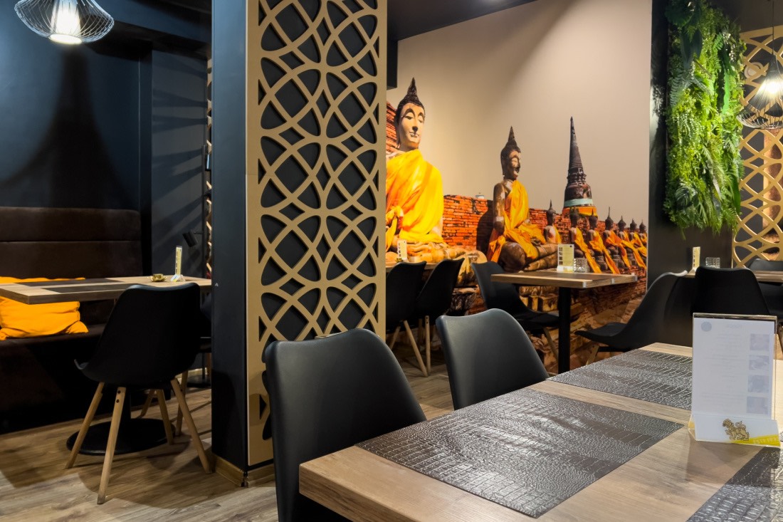 An den hellbraunen Holztischen vom Thai Orchid Restaurant stehen dunkelbraune Stühle. Im Hintergrund befinden sich ein großes Wandbild mit Tempel samt Buddhafiguren sowie eine grüne Hängepflanze.